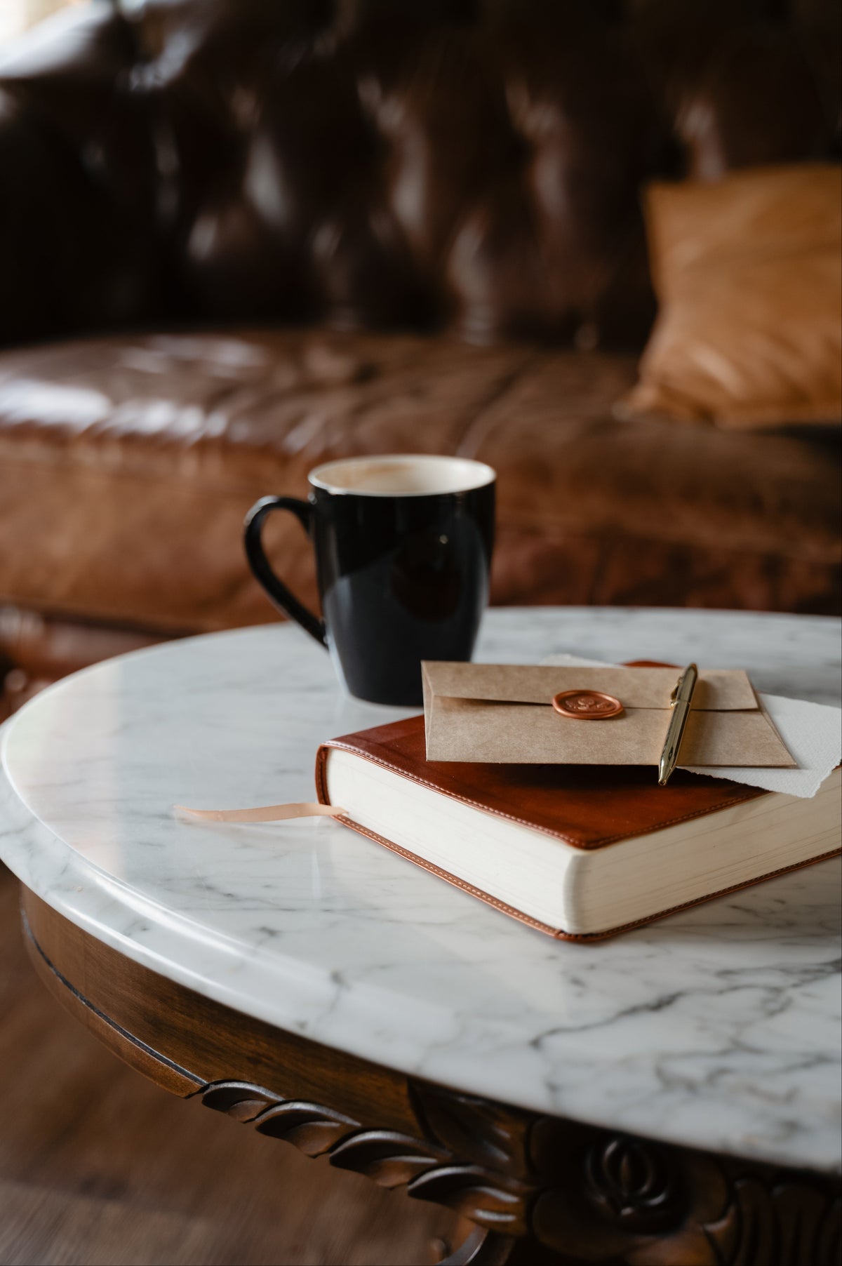 A styled coffee table scene with a black ceramic mug, a brown leather journal, and a kraft paper envelope with a wax seal, set against a blurred brown leather sofa background.