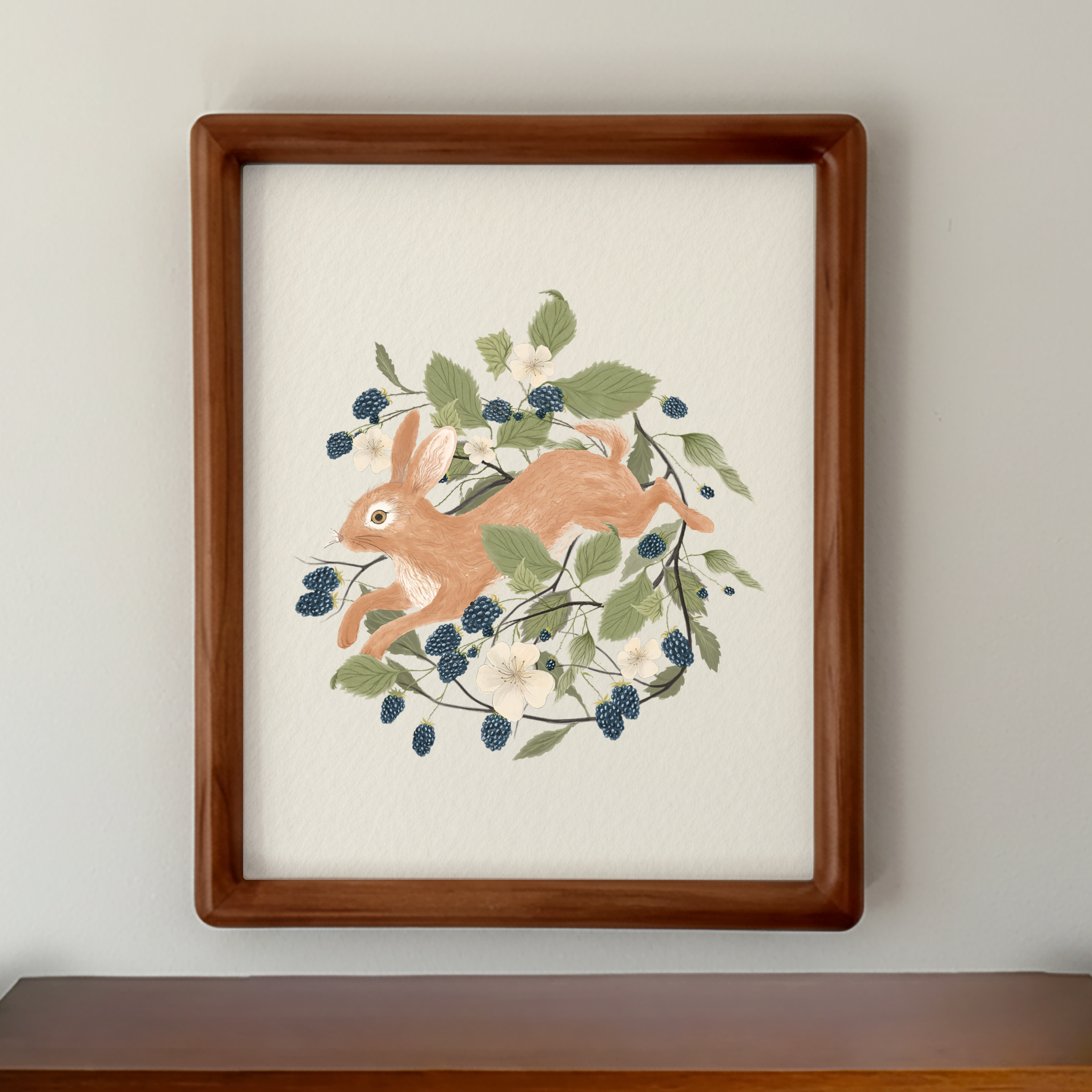 A framed art print featuring a watercolor-style illustration of a light brown rabbit surrounded by green leaves, dark blue berries, and small white flowers, set in a warm brown wooden frame against a light gray wall.